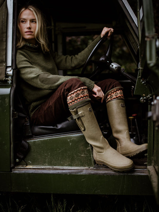 A woman in Campbells of Beauly Moleskin Plus Twos, with an olive sweater and tan boots, sits in the driver’s seat of a vintage green vehicle, hands on the wheel and looking toward the open door.